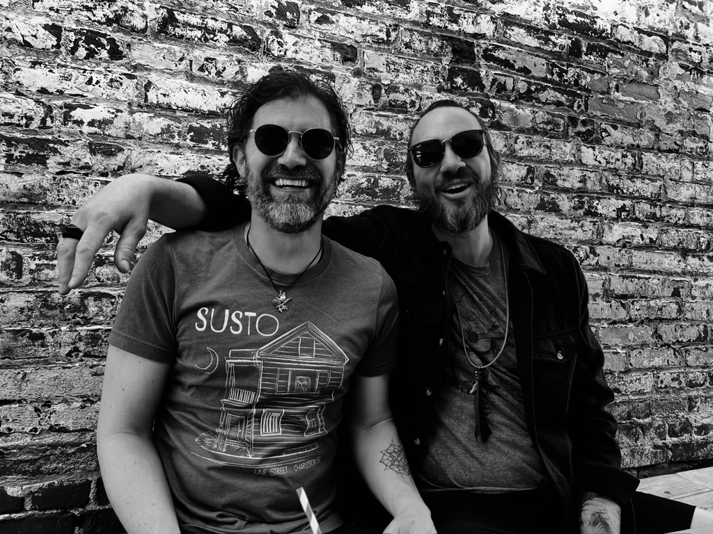 Michael and Doug in sunglasses, laughing against a brick wall, black and white